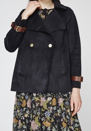 Veste noire double boutonnage en suédine synthétique avec un large col, des boutons dorés et des accents de brides en cuir brun sur les manches.