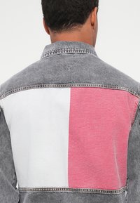 Denim jakna s leđima koja ima sivi dio, bijeli kvadrat i crveni kvadrat. Vidljivo šivanje i klasičan dizajn ovratnika.