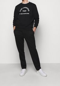 Uomo con indosso un sweatshirt nero di Karl Lagerfeld Paris, pantaloni neri e sneakers bianche, in piedi con le mani in tasca su uno sfondo bianco.