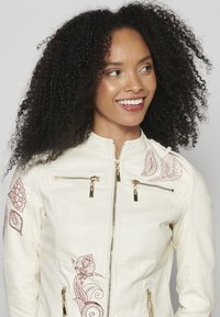 Veste en cuir blanc avec des zips dorés, ornée d'une broderie florale rouge complexe sur les épaules et les côtés, texture lisse, coupe ajustée.