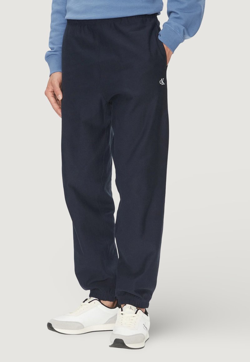 Homme portant un pantalon de jogging bleu marine avec des poignets élastiques, un sweat-shirt bleu clair, et des baskets blanches avec des détails noirs et gris.
