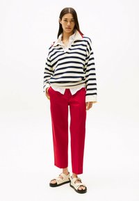 Pull en tricot rayé bleu marine et crème, col rouge, associé à un pantalon rouge et des sandales légères avec une semelle noire, reflétant un style décontracté.