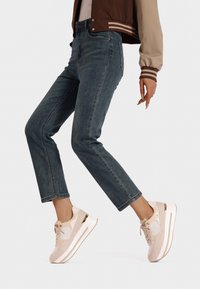 Sneakers rosa chiaro con tomaia testurizzata e rete bianca, caratterizzati da un dettaglio dorato sulla suola. Accoppiati con jeans scuri a gamba dritta.