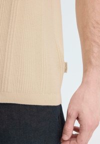 Beige gebreid top met een klein stoffen label en de rechterarm en hand van een persoon ernaast, die een donkere broek draagt.