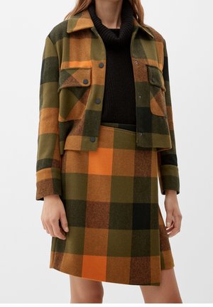 Femme portant une veste et une jupe assorties en laine à carreaux verts, orange et noirs, avec un col roulé noir en dessous.