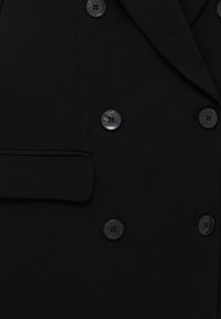 Blazer noir croisé à double boutonnage avec de gros boutons noirs brillants et une poche à rabat sur le côté gauche.