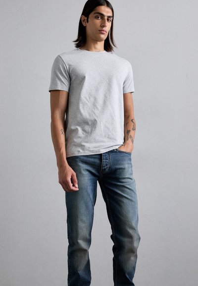 Jovem com cabelo escuro e longo, vestindo uma camisa cinza de mangas curtas e jeans azuis, de pé com uma mão no bolso contra um fundo liso.