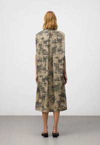 En person med medellångt hår vänder ryggen mot kameran, klädd i en mönstrad beige klänning och svarta klacksandaler med blockklackar, mot en enfärgad grå bakgrund.