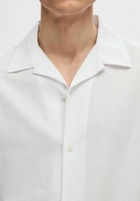 Camicia bianca in lino a maniche corte con colletto a punta, dotata di una patta con bottoni e una sottile trama del tessuto.