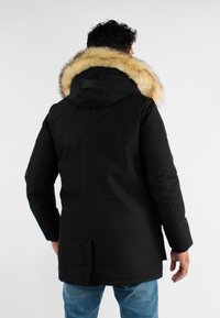 Parka noire avec une grande capuche en fausse fourrure, présentant une fermeture éclair, des poches latérales et une coupe ajustée pour un look fitted.