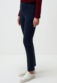 Pantalons bleu marine, coupe ajustée, avec poches avant, fermeture à bouton et texture lisse, associés à des baskets blanches.