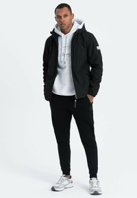 Veste à capuche noire sur un sweat-shirt gris avec cordons de serrage, associée à un pantalon de jogging noir et des baskets grises. Design simple et moderne.