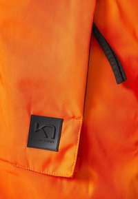 Tissu orange vif avec un patch carré noir comportant un logo stylisé « K » et le texte « KARITRAA » embossé.