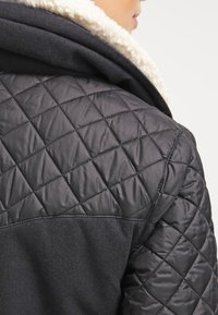 Veste matelassée noire avec un col en agneau texturé et une base en tissu lisse, présentant une couture diagonale à l'arrière.