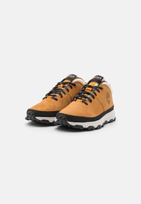 Zapatos de senderismo de ante marrón con cordones negros y suelas de goma. Presentan un logotipo en el lateral y una entresuela blanca en contraste. Patrón de tracción en las suelas.