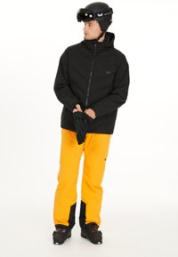 Svart isolerad jacka, gula byxor med svarta detaljer, svarta skidstövlar och handskar. Modellen bär en svart hjälm med visir.