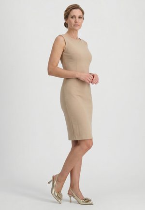 Žena stojí, má na sobě béžové přiléhavé šaty bez rukávů po kolena a zlaté slingbacky na podpatku s květinovou ozdobou.