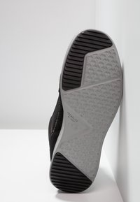 Černé tenisky s texturovanou šedou podrážkou, která má vzorovaný profil, a šitým černým svrškem. Označeno "TREADLITE by UGG."