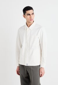 Cămașă albă cu mâneci lungi, cu guler, având un buzunar pe piept, dintr-un material de bumbac fin și cu o croială lejeră. Asortată cu pantaloni gri.