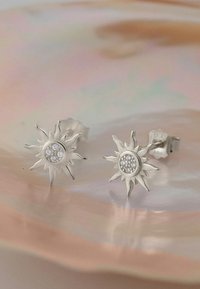 Silverörhängen i solform med en central cirkel av små klara stenar, som visar en matt yta och spetsiga strålar.