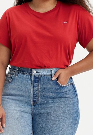 Vrouw draagt een rood Levi's T-shirt dat is ingestopt in een hooglopende, lichtblauwe spijkerbroek, met één hand in de zak en een armband om de linkerpols.