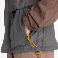Chaqueta ligera de tela gris y marrón, con puños elásticos, bolsillo oculto con cremallera amarilla y dobladillo ajustable.