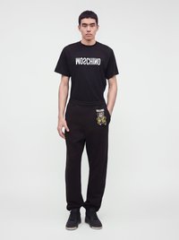 Млад мъж стои, носещ черна тениска Moschino с обърнат текст и черно спортно долнище с графика на плюшено мече на джоба.