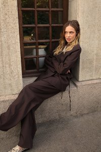 Long, loose-fitting brown trousers and matching oversized top with drawstring details. Model leans against a stone wall near a window.