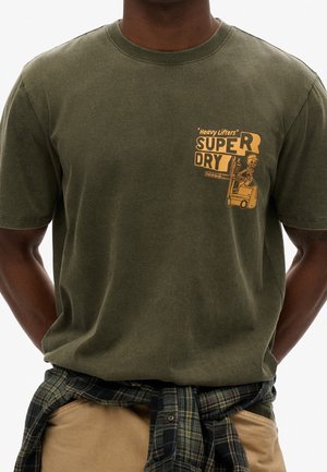 T-shirt en coton vert olive avec un imprimé graphique jaune, incluant un squelette et le texte « Super Dry » sur le côté gauche de la poitrine.
