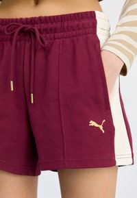 Shorts bordeaux avec une finition texturée, présentant un panneau latéral crème, une taille élastique et des éléments en métal doré. Logo affiché sur le devant.