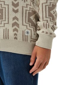 Beige und braun gemusterter Strickpullover mit einem kleinen "Honest Rules"-Label über dem gerippten Saum, getragen zu blauen Jeans.