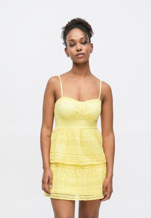 Robe en dentelle jaune avec un corsage ajusté, des bretelles fines et une jupe à étages. Le tissu présente des motifs complexes et une finition texturée.