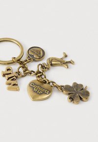Porte-clés en laiton avec un pendentif rond portant l'inscription "Carhartt WIP", un pendentif en forme de cœur, un trèfle à quatre feuilles, et une figurine, tous reliés par une chaîne.