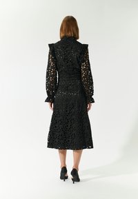 Schwarzes Midikleid aus Spitze mit floralen Ausschnitten, gerüschtem Kragen und langen Ärmeln. Kombiniert mit schwarzen Absätzen, hat eine taillierte Passform und einen fließenden Rock.