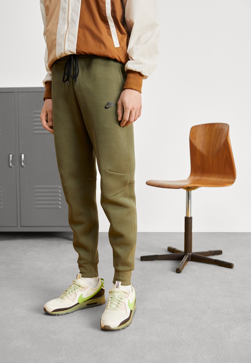 Olīvu zaļas Nike sporta bikses ar elastīgu jostasvietu un kabatām, apavi ar baltu un zaļu Nike dizainu, stāvot uz pelēka grīdas.