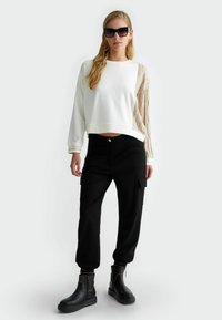 Felpa bianca corta con maniche beige texturizzate, abbinata a pantaloni cargo neri e stivaletti neri con cerniere. Il modello indossa occhiali da sole scuri.