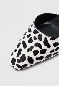 Mule cu vârf ascuțit, având o bază albă cu imprimeu leopard negru din material moale și texturat. Interior negru și design elegant.