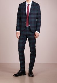 Traje a cuadros en verde oscuro y marino, ajustado. Presenta una chaqueta de corte sencillo, pantalones slim, camisa blanca y corbata burdeos. Zapatos de cuero negros.