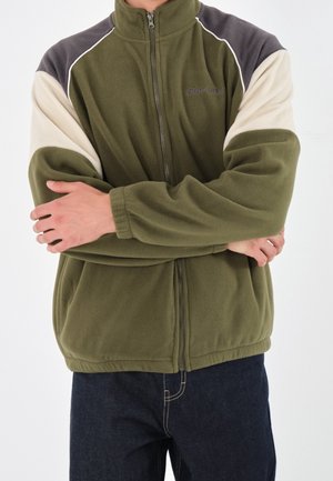 Veste zippée en polaire verte olive, avec des accents crème et gris foncé le long des épaules. Texture douce, avec des poignets et un ourlet élastiques.
