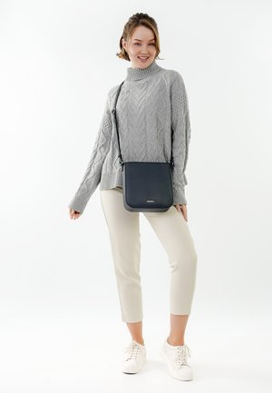 Mujer joven con suéter gris de punto trenzado, pantalones beige recortados, zapatillas blancas, sonriendo mientras está de pie con un bolso cruzado negro.