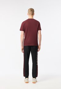 En kastanjefärgad t-shirt med kort ärm, kombinerad med svarta byxor som har en vit logga och förstärkta maroon sidostycken, visad bakifrån.