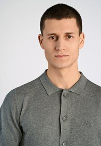 Grau gestricktes Poloshirt mit Kragen und drei Knöpfen, das eine subtile Textur und ein Logo auf der Brust aufweist.