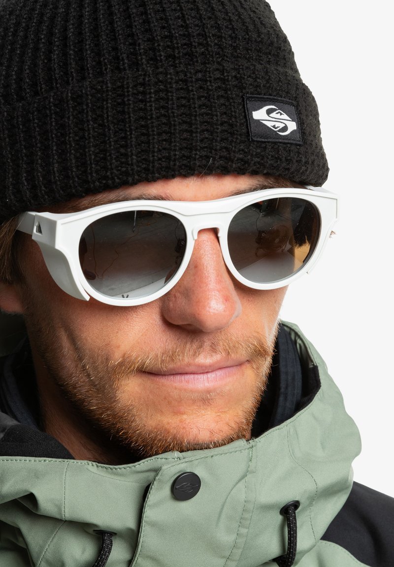 White sunglasses with rounded frames and dark lenses, worn with a black ribbed beanie and green jacket featuring a black accent.