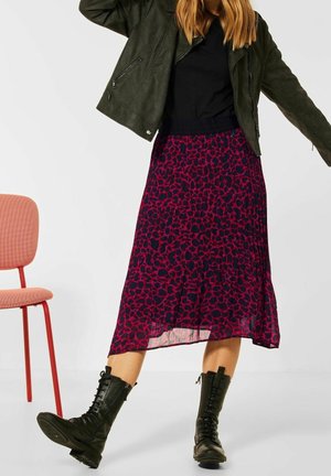 Falda maxi plisada con un estampado de leopardo púrpura y negro, combinada con un top negro ajustado y una chaqueta de cuero verde oscuro; llevada con botas de combate negras.
