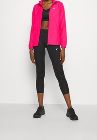 Sportliches Outfit mit einer leuchtend pinken, durchgehenden Reißverschlussjacke und schwarzen, eng anliegenden Leggings, ergänzt durch schwarze Sportschuhe.