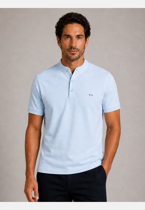 Mand med kort krøllet hår iført lyseblå kortærmet poloshirt med knappet stolpe og lille logo på brystet, står med hånden i lommen.