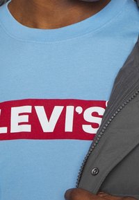 Jasnoniebieska koszulka z czerwonym prostokątem i białym napisem "LEVI’S", częściowo przykryta szarą kurtką z suwakiem z przodu i teksturowanym materiałem.