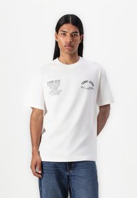 White cotton t-shirt with black printed text, short sleeves, and a round neck. Worn with blue denim jeans.