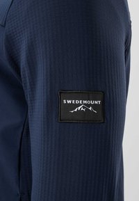Manche en tissu texturé bleu marine avec un patch rectangulaire noir affichant le texte "Swedemount" et un logo de montagne blanc.