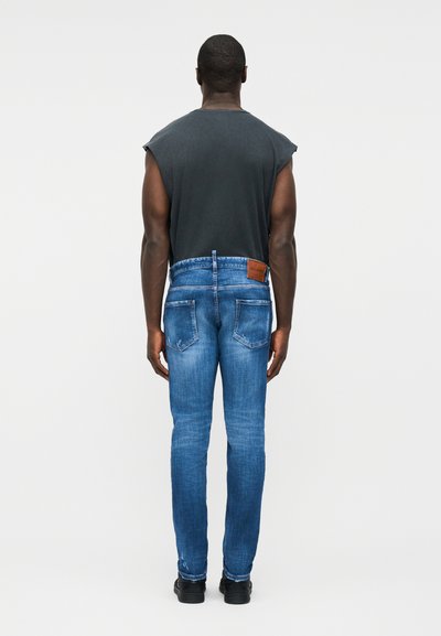 Homme portant un débardeur noir et un jean bleu, vu de dos sur un fond clair uni.